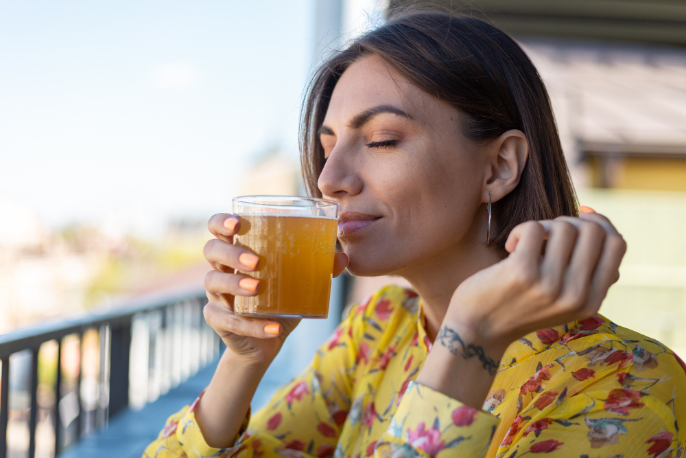 Kobieta w sukience w letniej kawiarni, pijąca chłodną szklankę kombuchy z piwem i wąchająca zapach piwa z zamkniętymi oczami