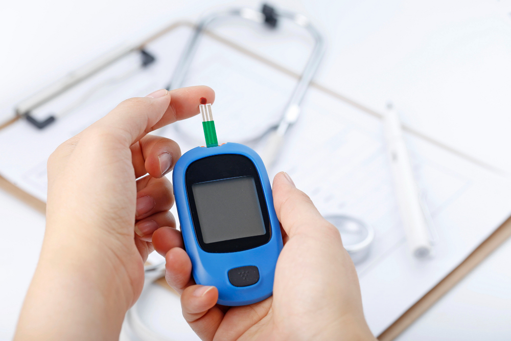 Dłoń trzymająca glukometr mierzący poziom cukru we krwi, tło to stetoskop i plik wykresu